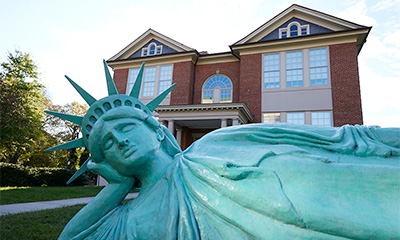 Replica of the Statue of Liberty lying on its side in the foreground, with a red-brick building and white-trimmed windows in the background.