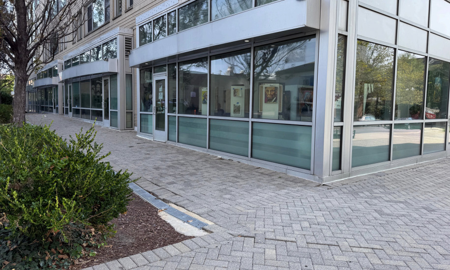 Modern glass-front building with large windows and artwork displayed inside, along a paved sidewalk lined with trees and landscaping.