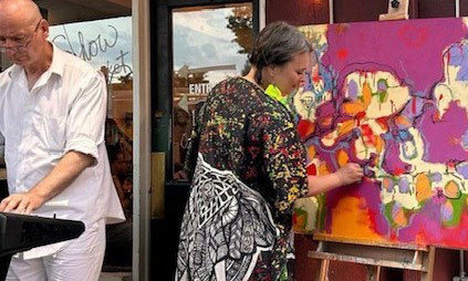 Person paints a colorful abstract artwork on an easel outside a building while another person stands at a nearby table.