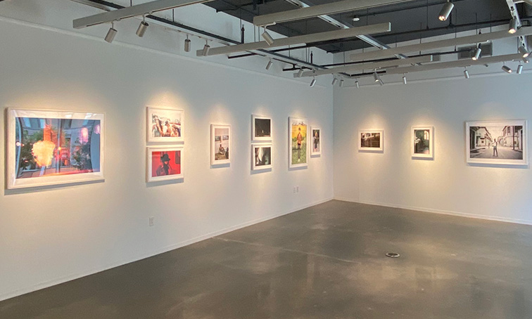 Bright, modern gallery with white walls displaying framed artworks and overhead track lighting in an open exhibition space.