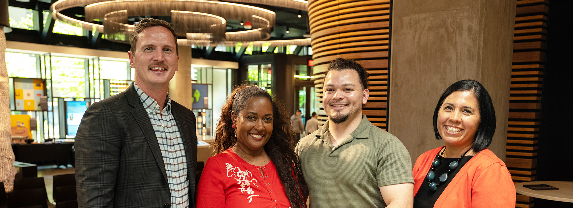 Four people standing side by side indoors in a modern building with wood columns, glass walls, and circular ceiling lights. Large windows and lobby seating are visible in the background.